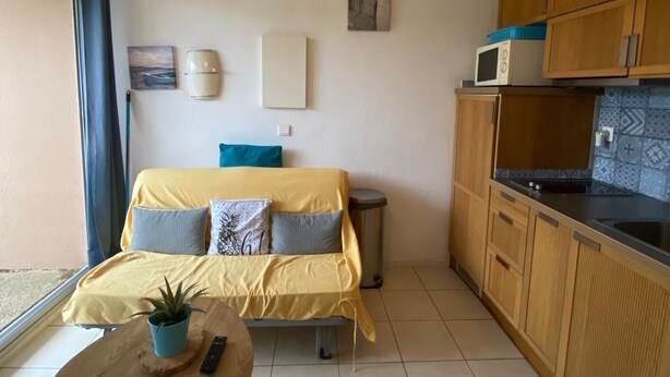 Ein kompakter Wohnraum mit Bett, kleinem Tisch mit Pflanze, Küchenzeile mit Holzschränken und einer Pendelleuchte.