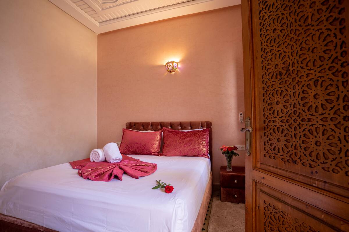 A hotel room with a neatly made bed, a decorative wall light, and a wooden door with intricate carvings.