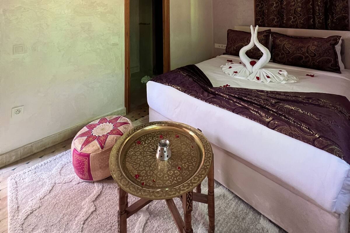 A hotel room with a bed, a television mounted on the wall, a decorative ceiling light, a small table with a tray, and a patterned rug.