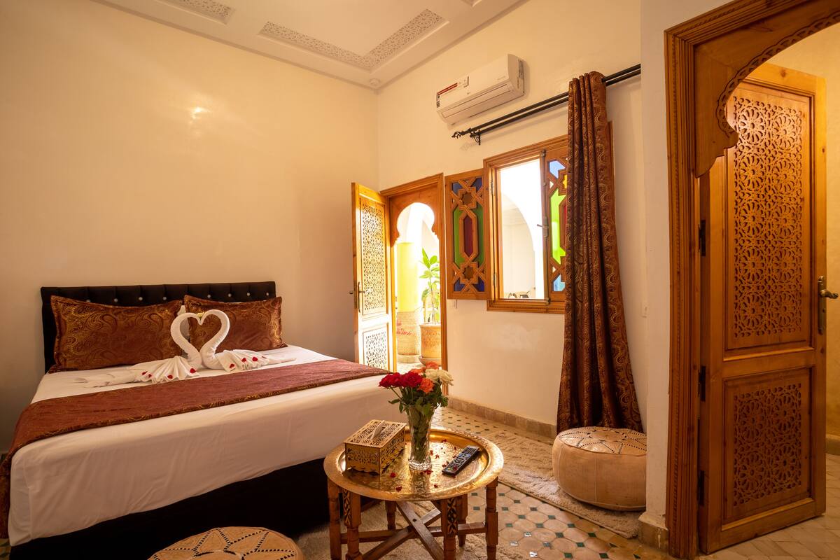 A hotel room with a bed, a small table with a vase of flowers, a remote control, and a decorative door.