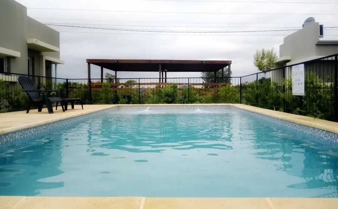 Ein rechteckiger Pool mit Fliesenrand, umgeben von einem niedrigen Zaun und einem überdachten Sitzbereich mit Stühlen.