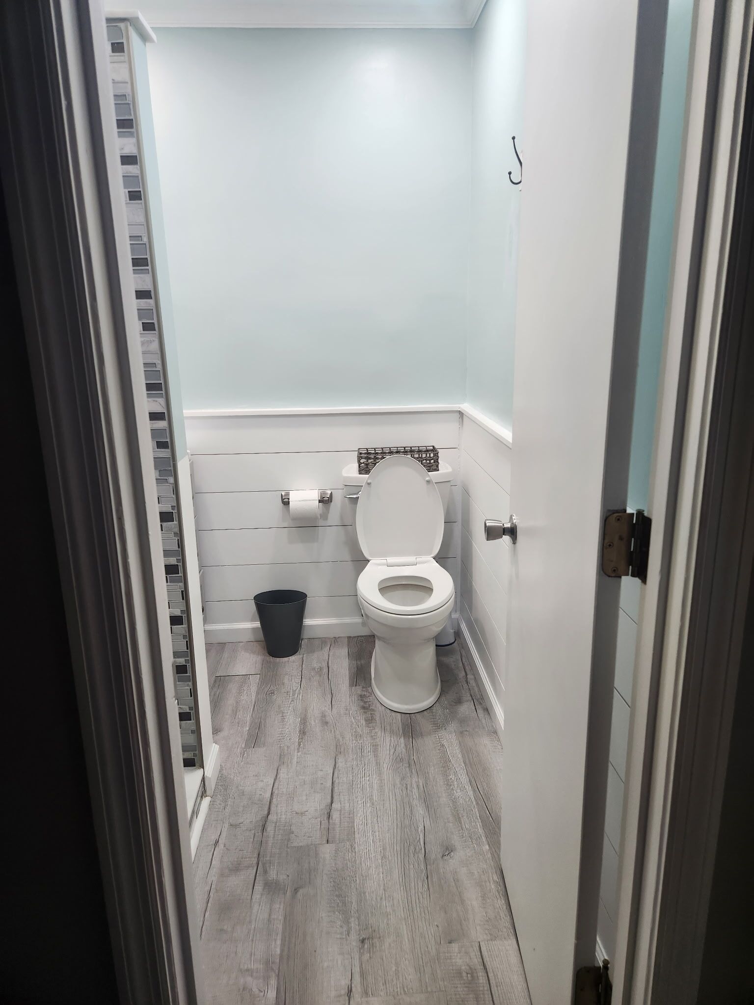 A small, narrow bathroom with a white toilet, a black trash bin, and a wooden floor.