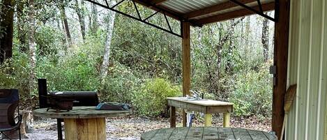 Un espace extérieur abrité comprenant deux tables en bois, un seau noir portant l’inscription « UNIQUEMENT POUR LES BOÎTES DE CONSERVE », et un barbecue.