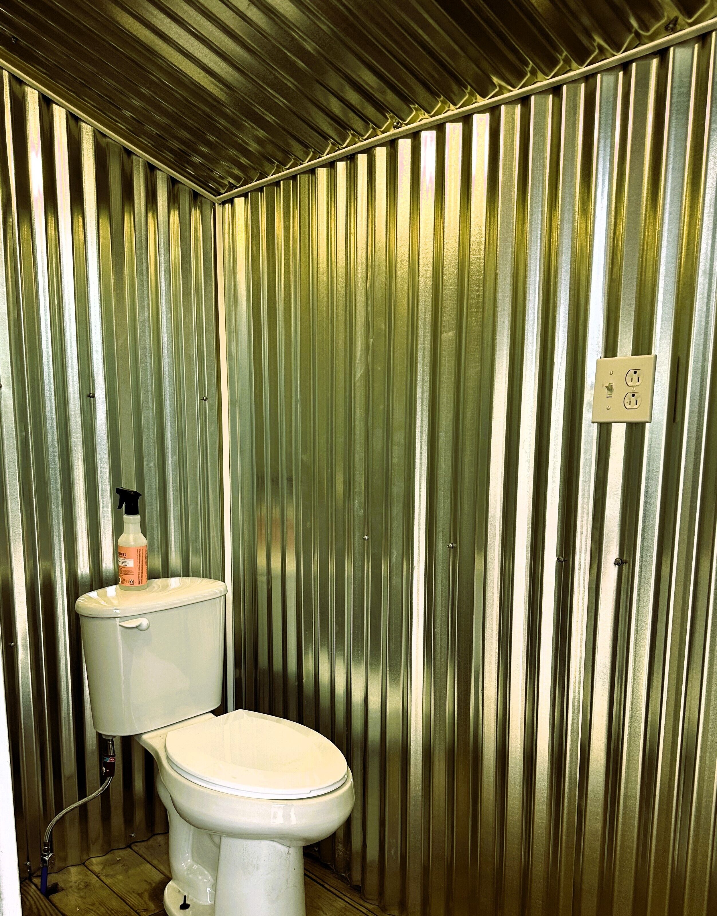 A small, enclosed bathroom with a toilet and a sink, featuring a corrugated metal wall and a visible electrical outlet.