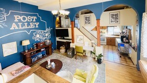 Una sala de estar con pared azul, televisor, mesa de comedor y zona de cocina visible a través de un arco.