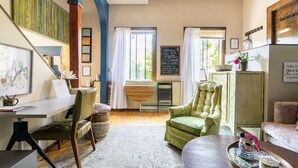 Una sala de estar acogedora con un escritorio, un sillón verde y un sofá. Hay una pizarra con un mensaje de bienvenida, una ventana con cortinas y un armario de madera.
