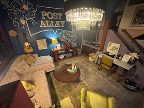 Una sala de estar acogedora con un sofá, una mesa de centro y una guitarra. Un candelabro cuelga del techo, y un mural en la pared dice 'POST ALLEY'.