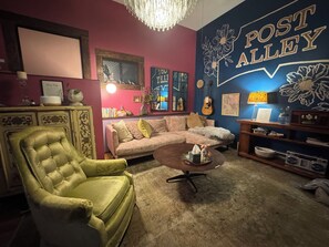A cozy living room with a green armchair, a beige sofa, and a coffee table. A wall mural reads 'POST ALLEY' and there are guitars and books on shelves.