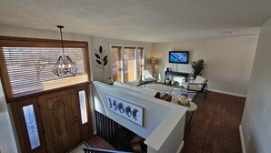 A modern living room with a white sofa, a coffee table, and a TV mounted on the wall. There is a staircase leading to an upper floor with wooden doors and a decorative leaf wall art piece. - Beautiful home just outside of Ogden Canyon (Ogden)