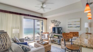 A living room with a large window overlooking the ocean, a ceiling fan, and a kitchen area with a bar stool and a television.