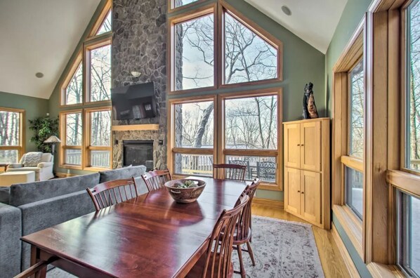 Un comedor amplio con una gran mesa de madera y sillas, una chimenea de piedra y ventanas grandes que ofrecen vistas al exterior.