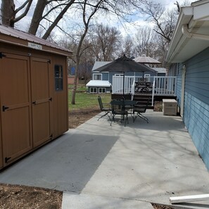 A covered patio area with a table and chairs, a shed labeled "HAPPY PLACE," and a blue house in the background. - Family Fun - 2 blocks from everything! (Arnolds Park)