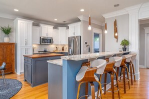 Cocina moderna con armarios blancos, electrodomésticos de acero inoxidable, una isla central con taburetes y luces colgantes.