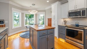 A modern kitchen with white cabinets, a wooden island, stainless steel appliances, and large windows offering a view of the outdoors.