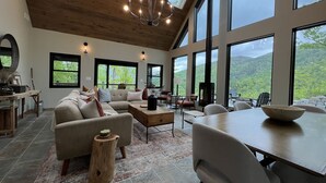 A spacious living room with a large sectional sofa, a dining table, and a fireplace. The room features large windows offering a view of the outdoors. - Scenic 7-acre mountain home, seasonal waterfall view, EV charger, dog friendly! (Marion)