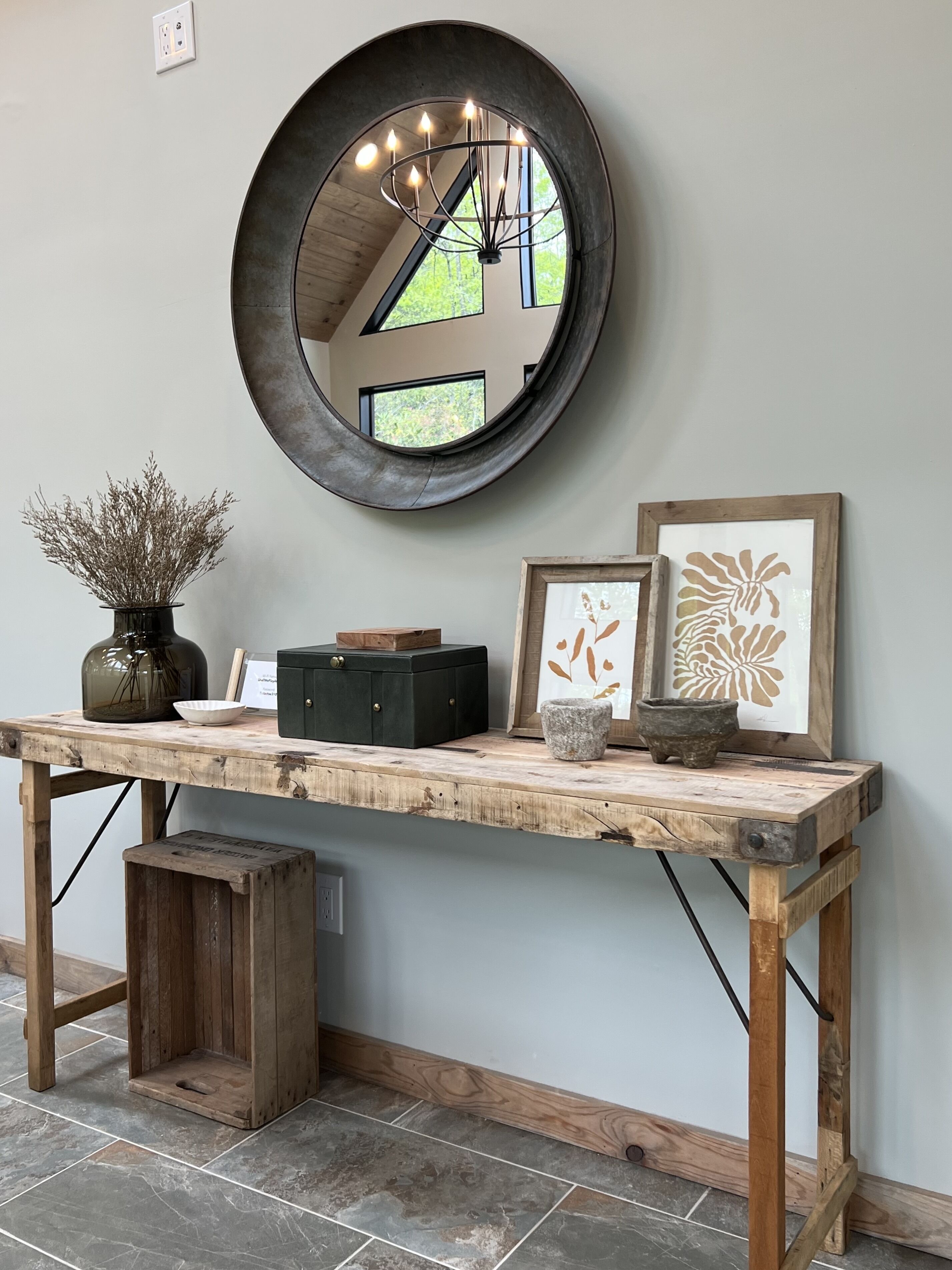 A rustic wooden desk with a mirror, a vase with dried plants, and framed artwork against a plain wall.