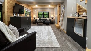A modern living room with a sofa, a coffee table, and a TV on a wooden cabinet. There is a window with a view of trees and a staircase leading to an upper level. - Scenic 7-acre mountain home, seasonal waterfall view, EV charger, dog friendly! (Marion)