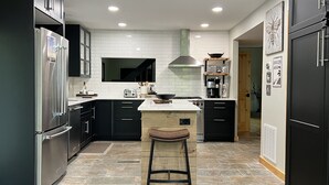 A modern kitchen with dark cabinetry, a stainless steel refrigerator, a central island with stools, and a large window above the sink. - Scenic 7-acre mountain home, seasonal waterfall view, EV charger, dog friendly! (Marion)