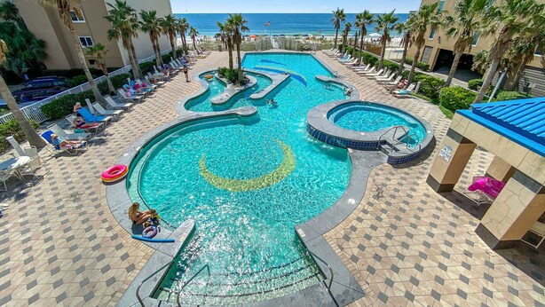 A swimming pool with a unique design, surrounded by lounge chairs and palm trees, with a view of the ocean. - Crystal Tower Unit 1508 | Snowbird Friendly | Indoor, Outdoor Pools, Hot Tub, Sauna, Gym (Gulf Shores)