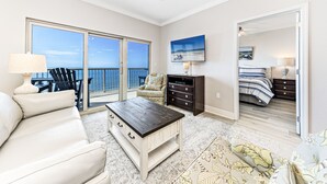 A spacious living room with a large window, a ceiling fan, a white sofa, a coffee table, and a view of the ocean. - Crystal Tower Unit 1508 | Snowbird Friendly | Indoor, Outdoor Pools, Hot Tub, Sauna, Gym (Gulf Shores)