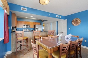 A dining area with a wooden table and chairs, a kitchen in the background, and a blue wall.