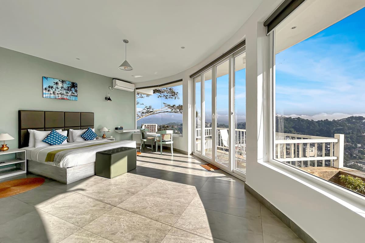 A modern bedroom with a large bed, a desk, and a balcony with a city view.