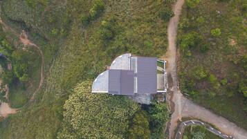 An aerial view of a building with a curved roof, surrounded by dense greenery and a dirt path.