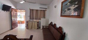 Sala de estar com sofá, televisão, quadro, área de cozinha com geladeira e porta que dá para área externa.