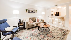 A living room with a sofa, armchairs, a coffee table, and a rug. There are two lamps, a painting, and a small table with a basket.