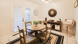 A dining area with a round wooden table, four chairs, a small potted plant, a wall clock, a mirror, and a bar area with a stool.