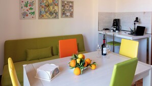 Ein Essbereich mit einem weißen Tisch, grün-gelben Stühlen, einem grünen Sofa und einer Küchenzeile mit einer Kaffeemaschine.