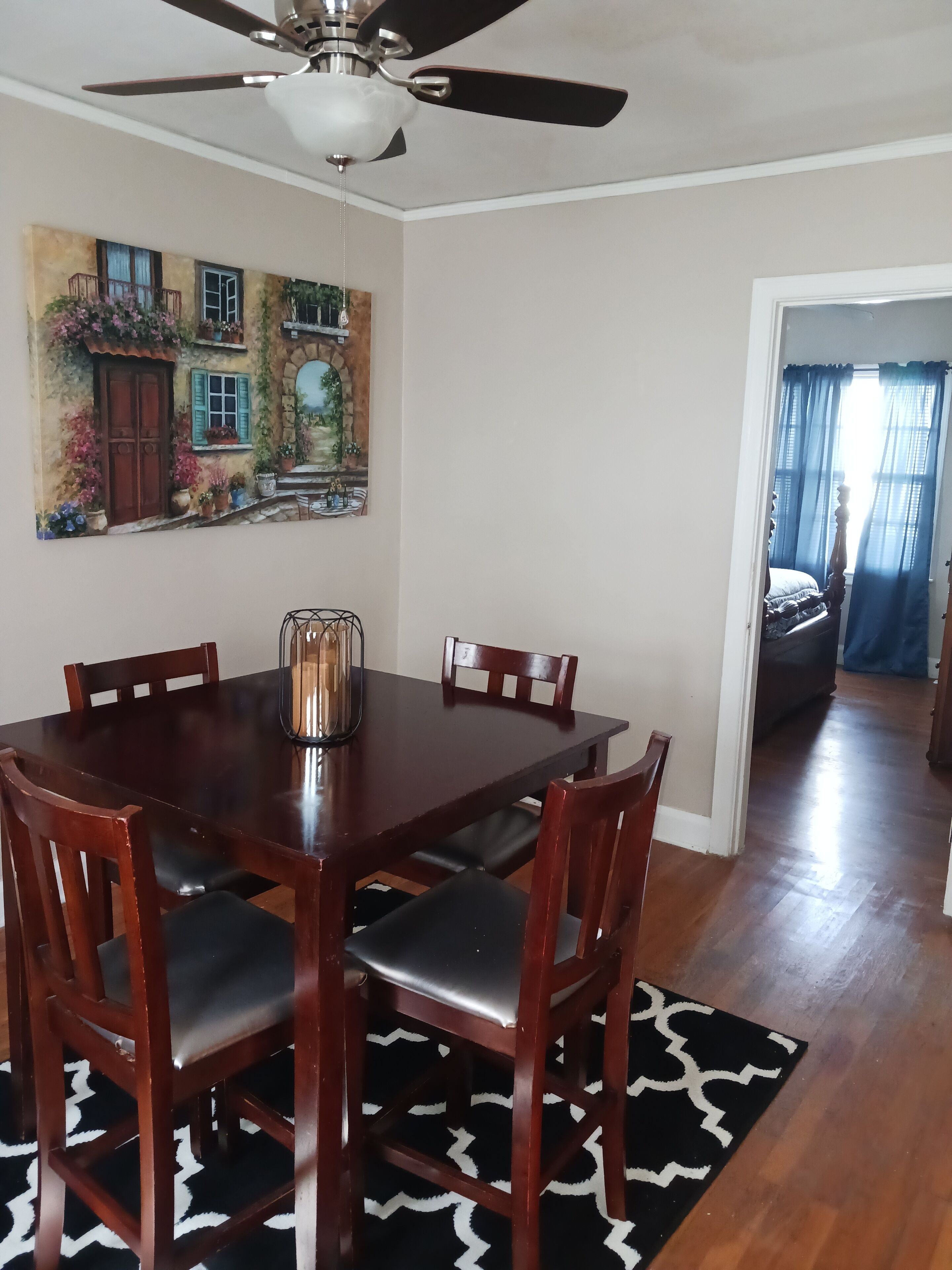 A dining area with a wooden table and chairs, a ceiling fan, and a painting of a house on the wall.