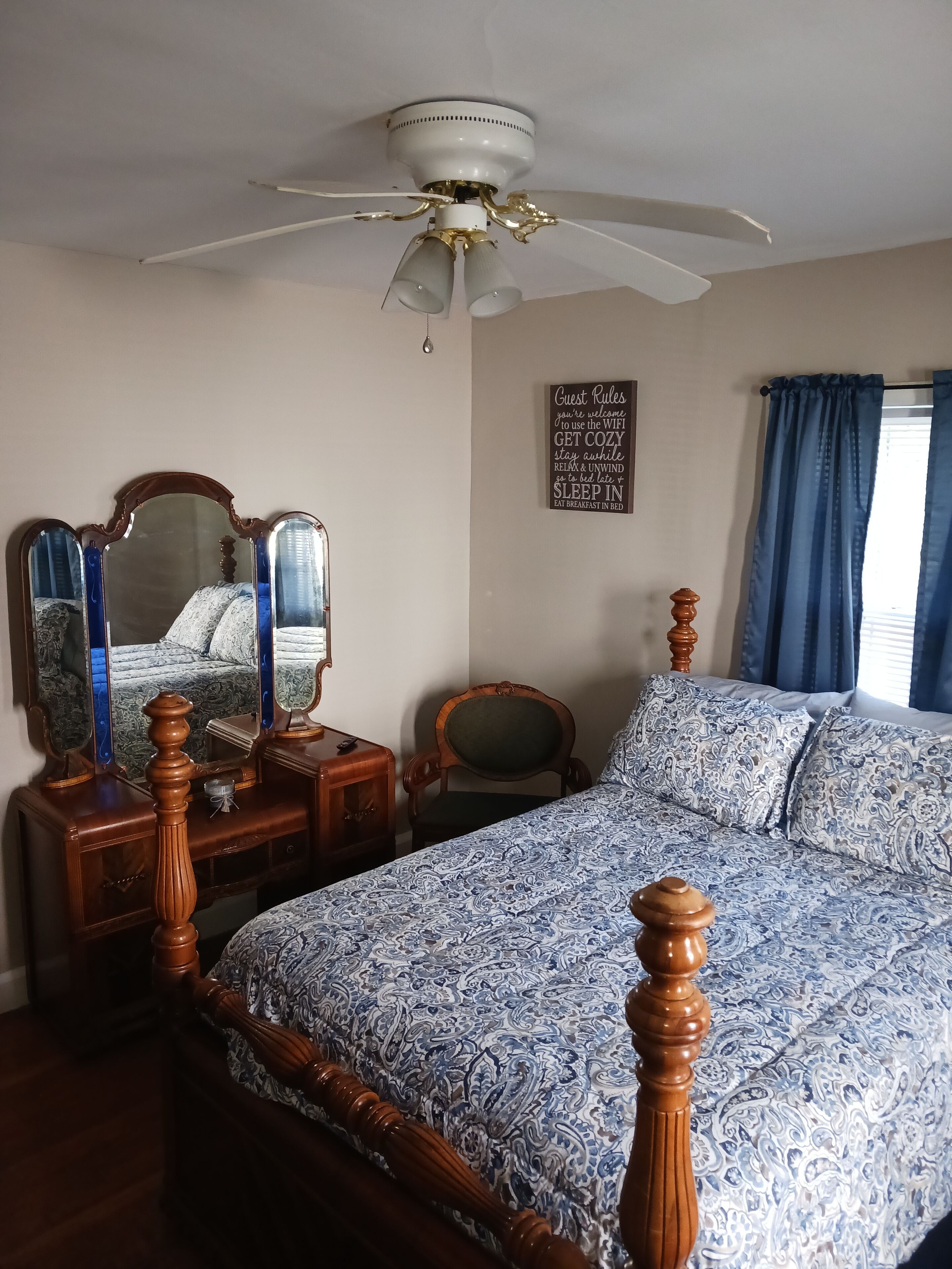 Een slaapkamer met een bed, een houten hoofdbord, een plafondventilator, een dressoir, een spiegel, een stoel, een bord aan de muur en een raam met gordijnen.