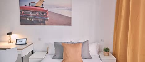 Habitación de hotel con cama, escritorio con lámpara, una planta pequeña y un cuadro con motivos playeros en la pared.