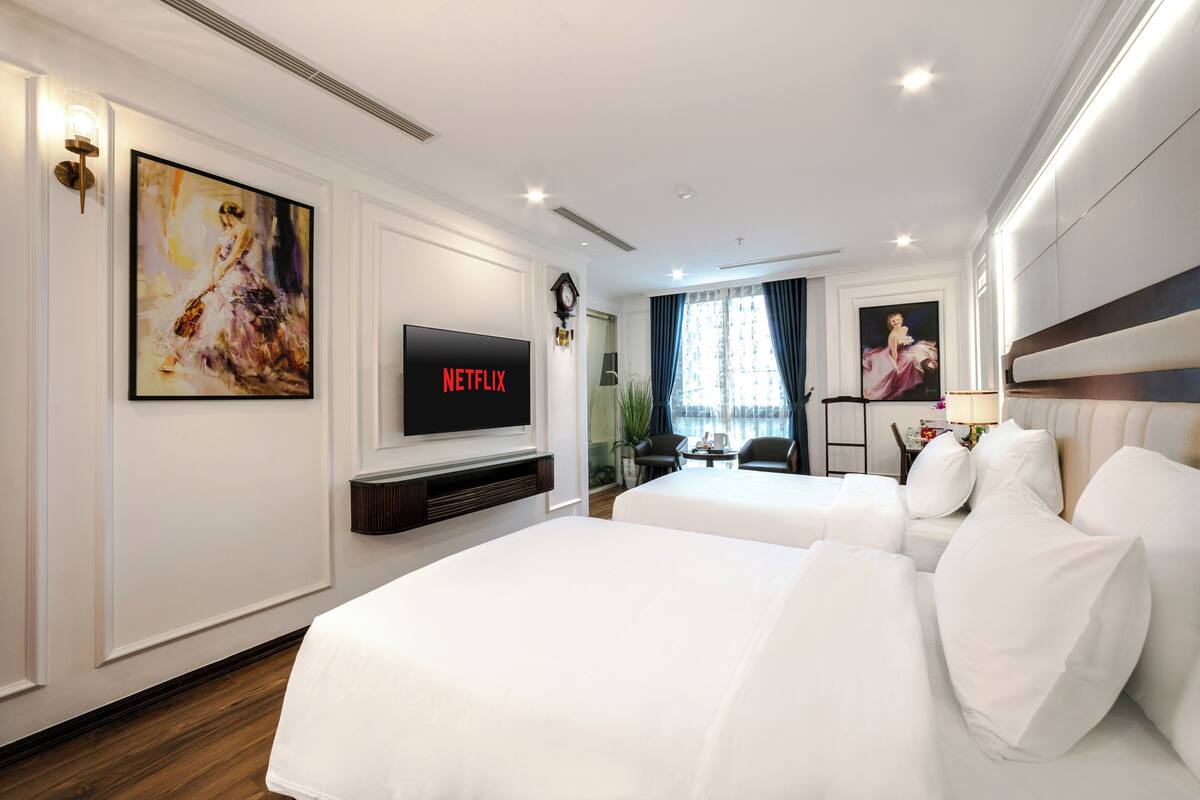 A hotel room with two beds, a TV, artwork, and a desk with a chair.