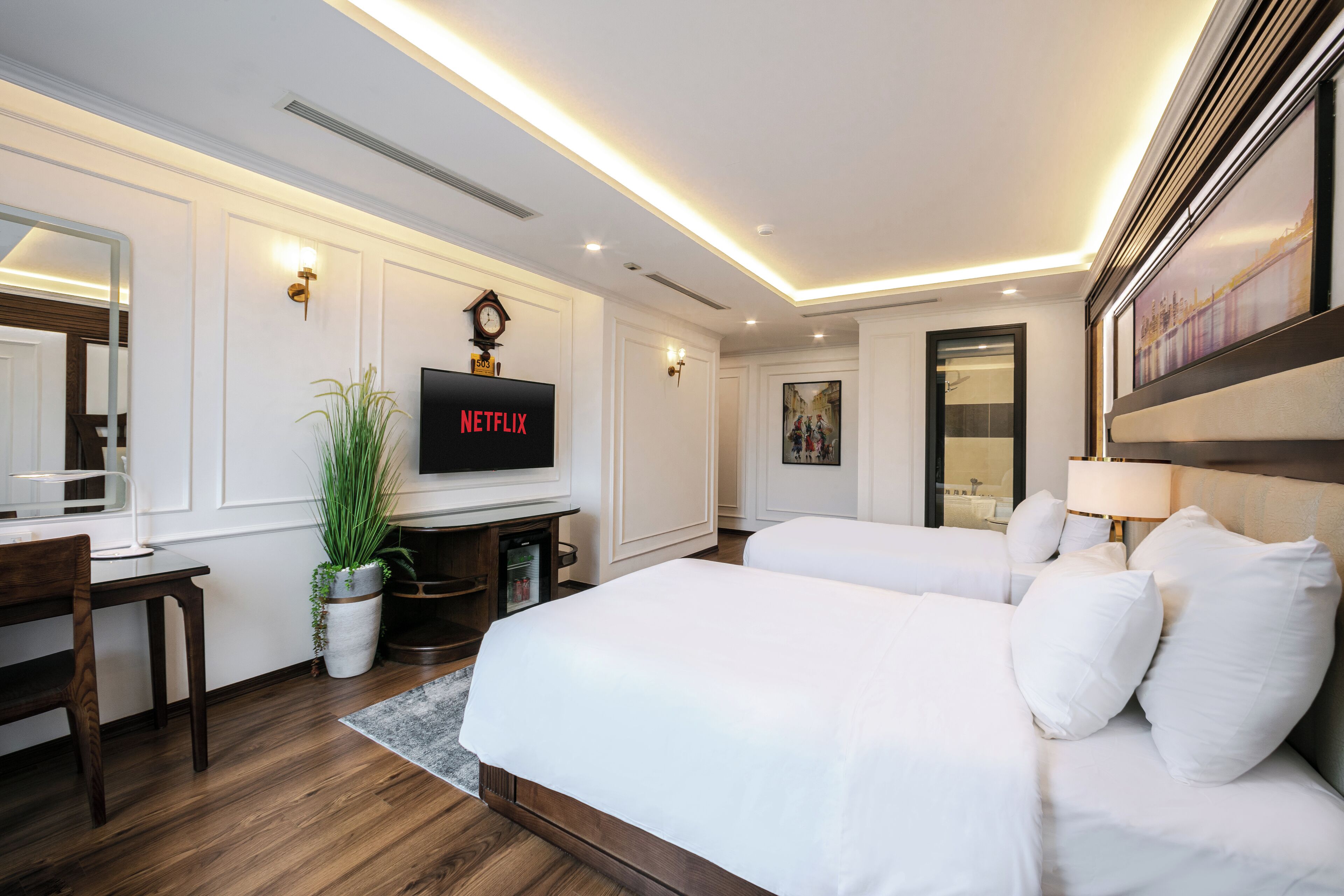 Quarto de hotel com televisão, duas camas, escrivaninha, planta e quadro na parede.