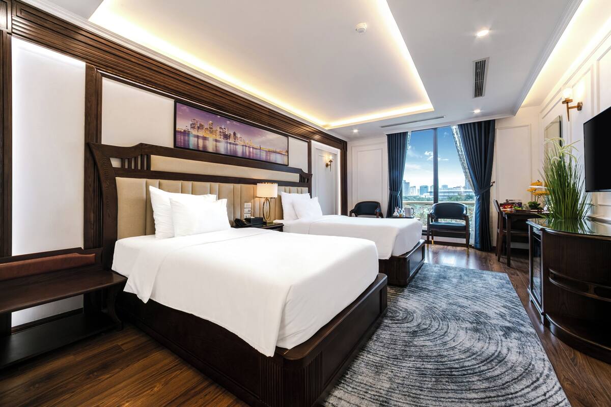 A hotel room with two beds, a large window with a city view, a flat-screen TV, and a desk with a chair.