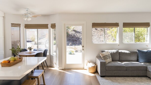 Un salon moderne avec un canapé gris, un coin repas avec une table et des chaises, et une cuisine dotée d’un comptoir blanc et de tabourets en bois.