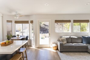 Un salon moderne avec un canapé gris, un coin repas avec une table et des chaises, et une cuisine dotée d’un comptoir blanc et de tabourets en bois.