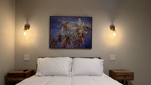 A bedroom with a large bed, two bedside tables, a wall-mounted painting of palm trees, and two wall-mounted lights.
