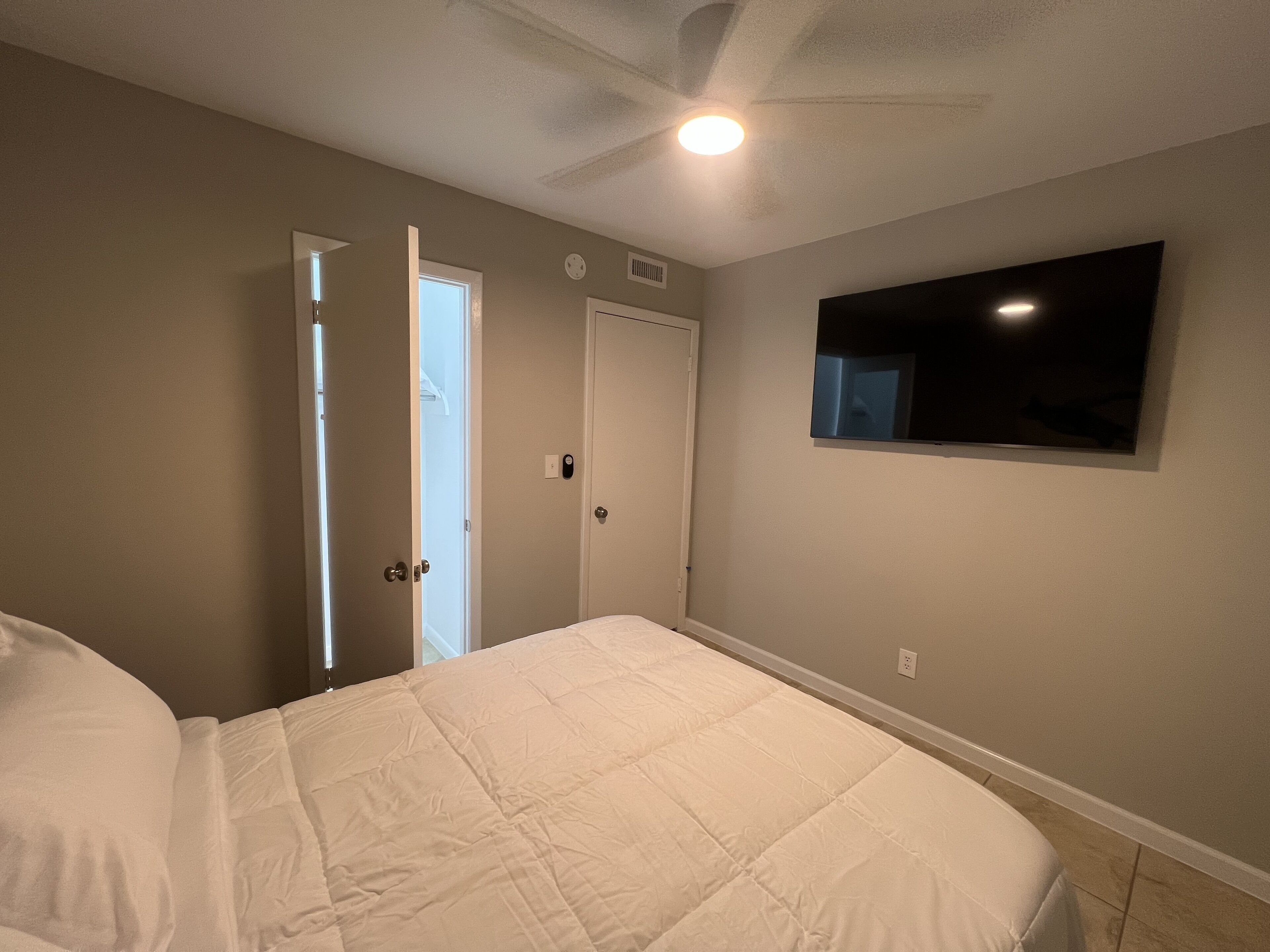 A bedroom with a bed, a ceiling fan, a TV mounted on the wall, and a door leading to another room.