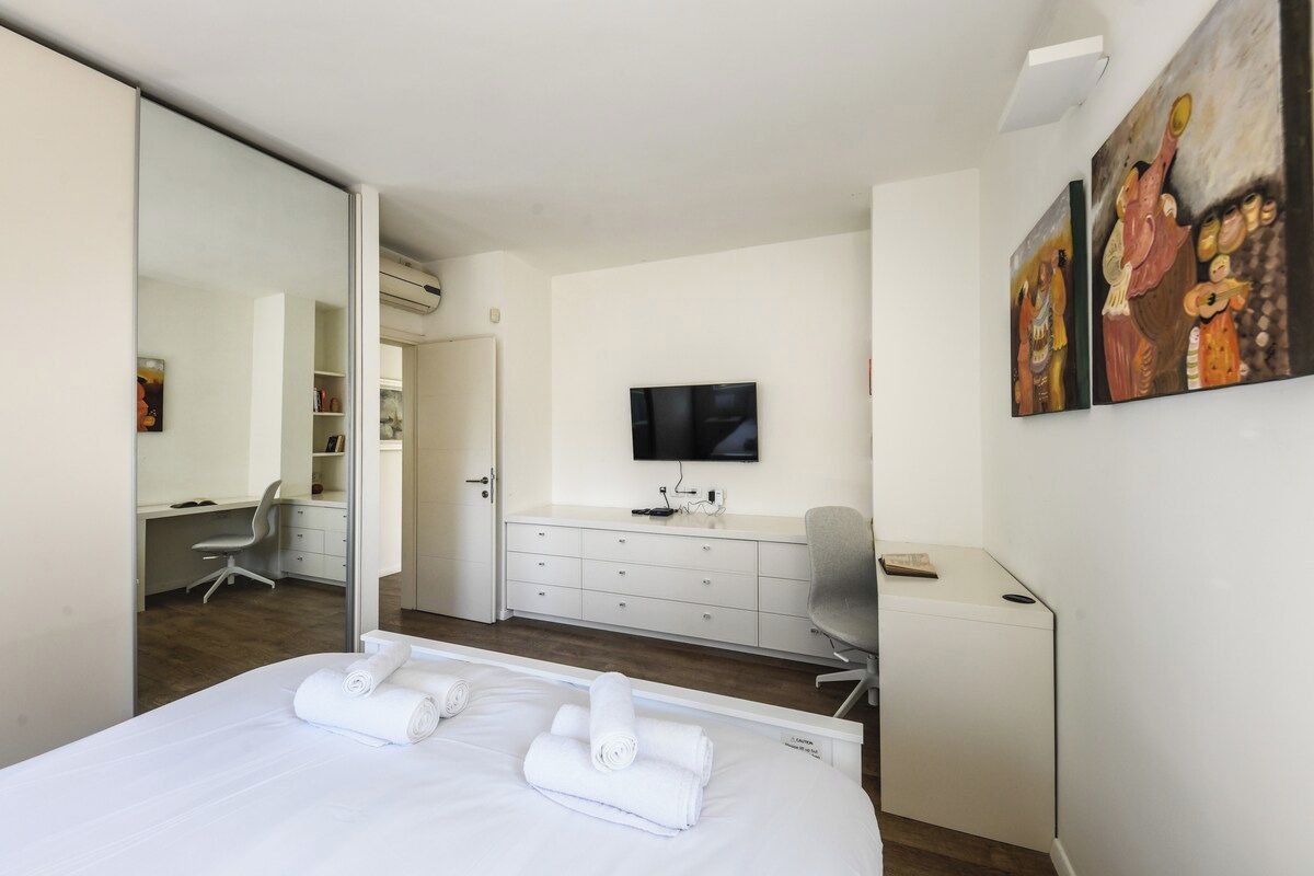 Quarto de hotel com cama, escrivaninha, cadeira, televisão e obras de arte na parede.