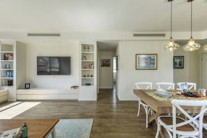 A modern living room with a flat-screen TV, white shelving, a dining area with a wooden table and white chairs, and pendant lighting.