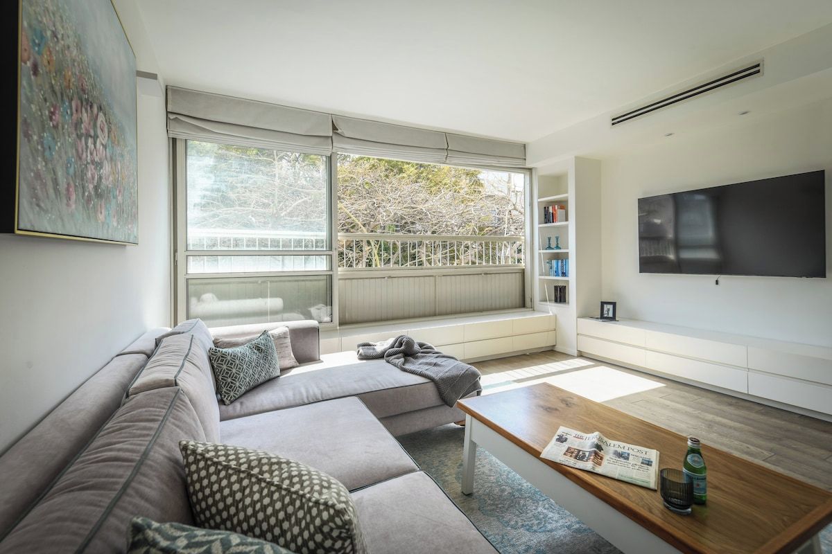 Een moderne woonkamer met een hoekbank, een salontafel, een flatscreen televisie en een groot raam met uitzicht op bomen.