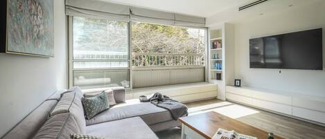 A modern living room with a sectional sofa, a coffee table, a flat-screen TV, and a large window with a view of trees.