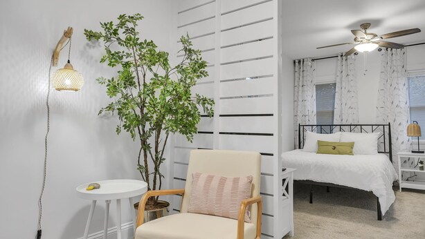 Un dormitorio con una cama, una silla, una mesita, una planta en maceta y un ventilador de techo.