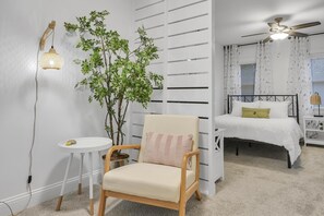 Un dormitorio con una cama, una silla, una mesita, una planta en maceta y un ventilador de techo.