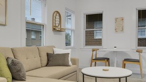 Un salón con un sofá color beige, dos sillas de madera, una mesita blanca y un espejo colgado en la pared.