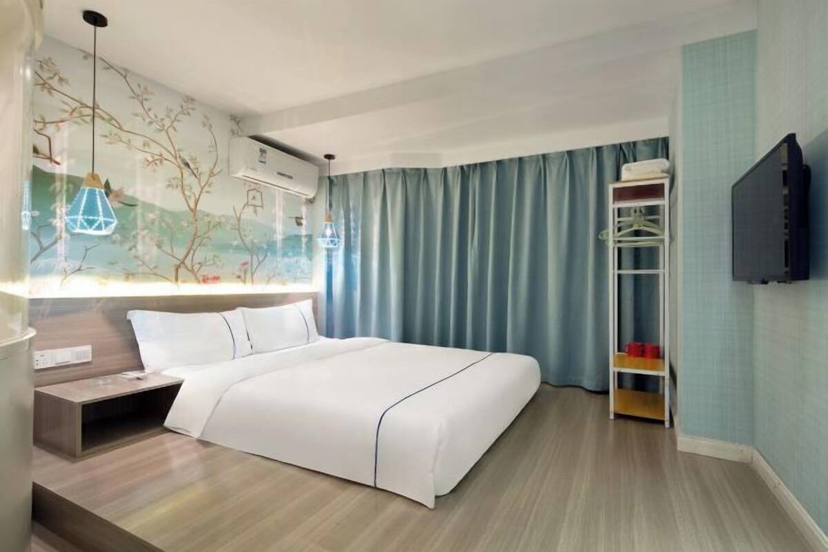 Una habitación de hotel con cama, mesita de noche, televisor, armario y ventana con cortinas.