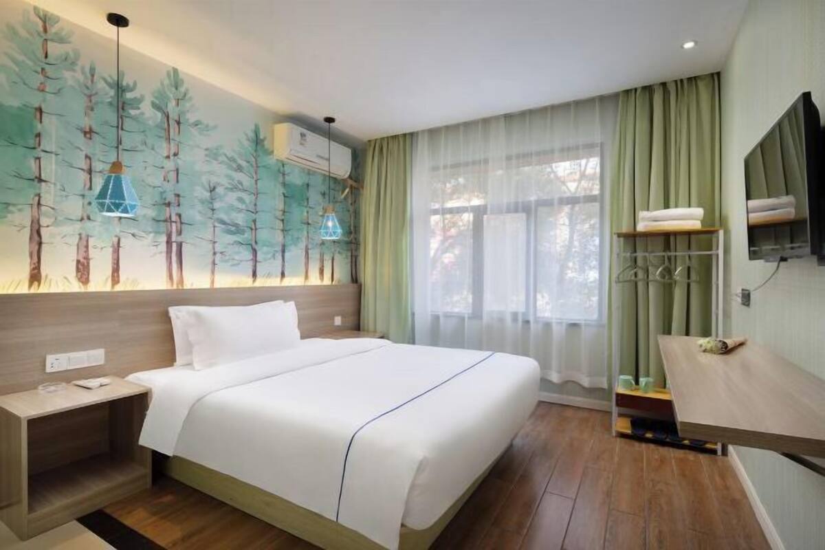 Habitación de hotel con cama, mesita de noche, televisor montado en la pared, ventana con cortinas translúcidas y estante con toallas.